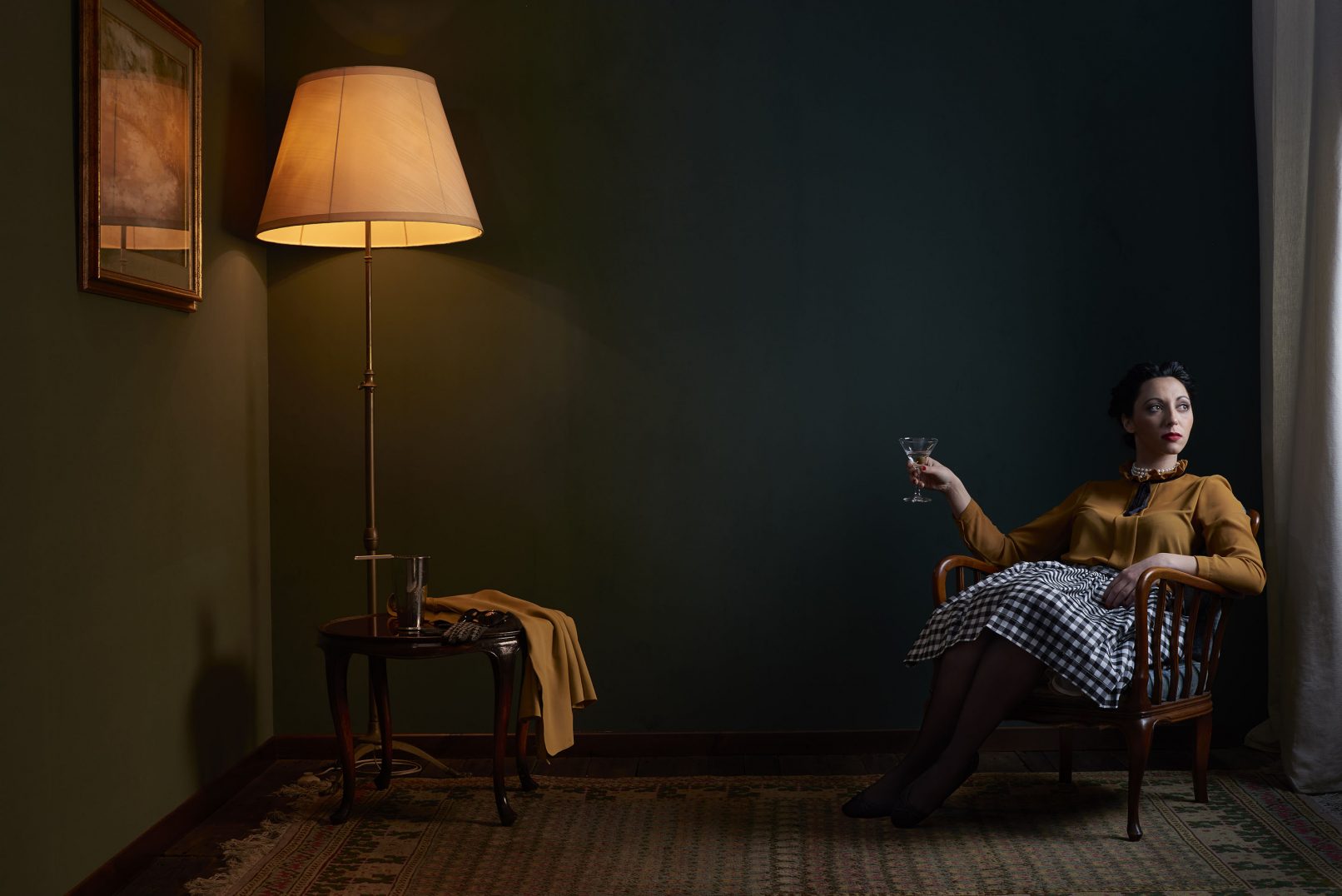 Historic portrait of a woman in 1950’s clothing set in a room with lamp, coffee-table, carpet and green walls. Set design according to 1950’s interior style.. Personal project dedicated to historic portraits. Image series by David Umberto Zappa - portrait, commercial and advertising photographer. Studio photography. Set-design. Ritratto storico di donna in abiti anni 50’ in stanza allestita secondo lo stile dell’epoca. Lampada, tappeto, pareti verdi, tavolino da fumo. La donna regge un bicchiere da Martini mentre guarda fuori dalla finestra. Progetto personale del fotografo commerciale, pubblicitario, ritrattista ed editoriale David Umberto Zappa. Fotografia in studio. Set-design. Fotografia con luce artificiale.