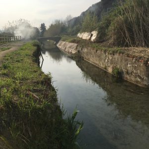 Canale Lunense nei prossi di Località Nave a Sarzana. Pista ciclabile pedonale sul lato sinitro. Vegetazione fitta sul lato destro e fumo sullo sfondo.