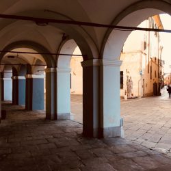 Piazza Matteotti Sarzana con facciate di palazzi storici e portici sul lato sinistro. Inferiate e portoni. Location Scout duzimage